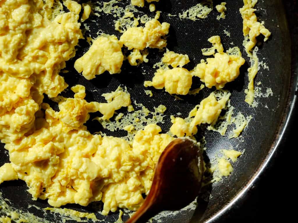 Scrambled vegan eggs cooking in a non-stick pan, with a wooden spoon resting on the side. The eggs are partially cooked, showing a creamy texture with some golden-brown cooked edges.